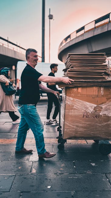 A man wearing a black shirt and blue jeans is pushing a large trolley loaded with cardboard boxes inside an urban outdoor setting, possibly part of a house or furniture removal process. The boxes are